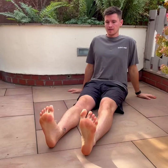 Man sitting on a patio with plants and a white wall in the background, doing foot strength exercises