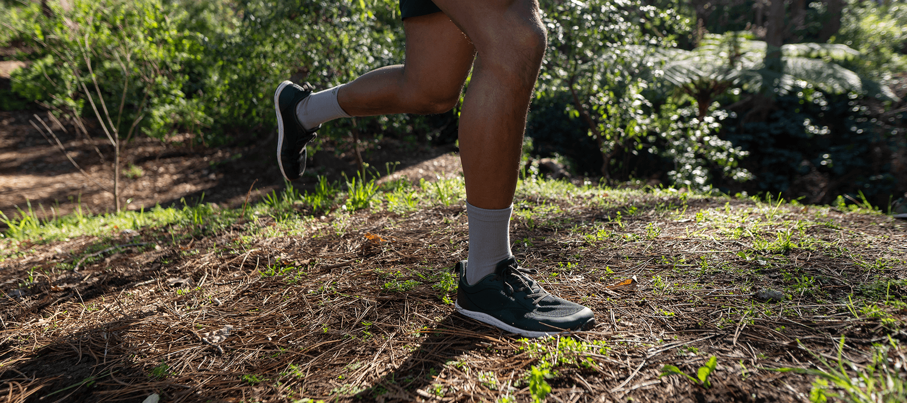 A close-up of a person's legs running outdoors on a grassy, pine-covered path, wearing Bahé Revive Flex Midnight. 