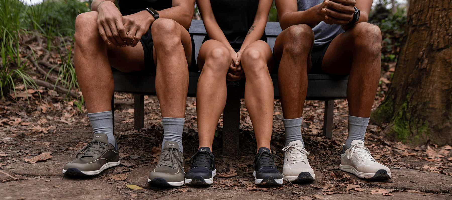 Three people sitting on a bench outdoors, showing their legs from the knees down in Bahé, socks, and running shoes on a wooded path.