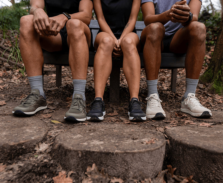 Three people sitting on a bench outdoors, showing their legs from the knees down in Bahé, socks, and running shoes on a wooded path.