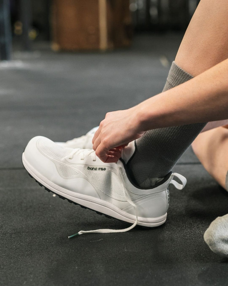 Person wearing a white sneaker with 'bahé' branding on a dark floor.