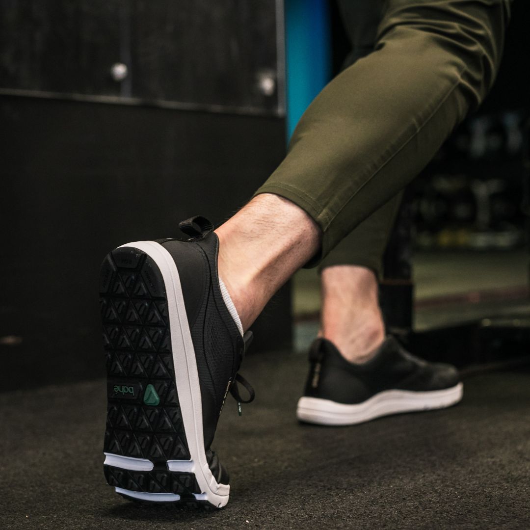 Person wearing black sneakers with white soles on a dark floor.